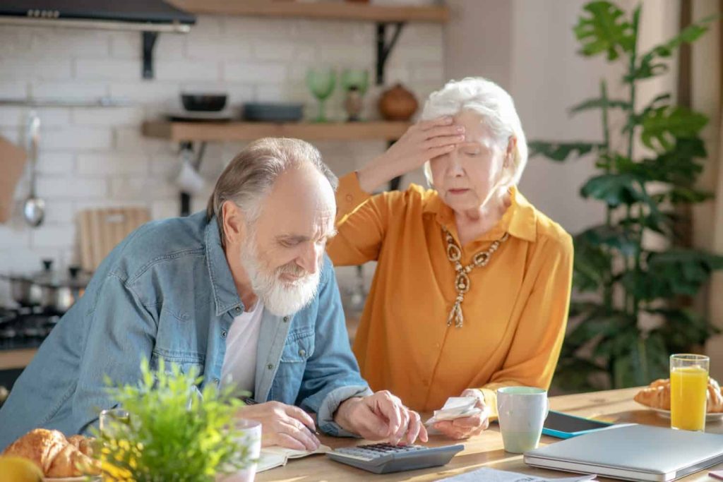 An elderly man with a white beard in a denim shirt sits at a kitchen table, looking at papers and using a calculator, while an elderly woman with white hair in a yellow shirt stands behind him, holding her forehead with a worried expression. On the table are a plant, croissants, orange juice, and a coffee mug.