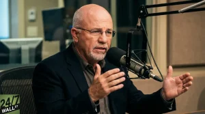 A bald man with a short white beard and glasses, wearing a dark suit jacket and dark shirt, speaks intently into a professional studio microphone. His hands are raised and gesturing as he talks, suggesting an engaging discussion. The background is blurred, showing office or studio equipment, and a '24/7 WALL ST' logo is visible in the bottom left corner.