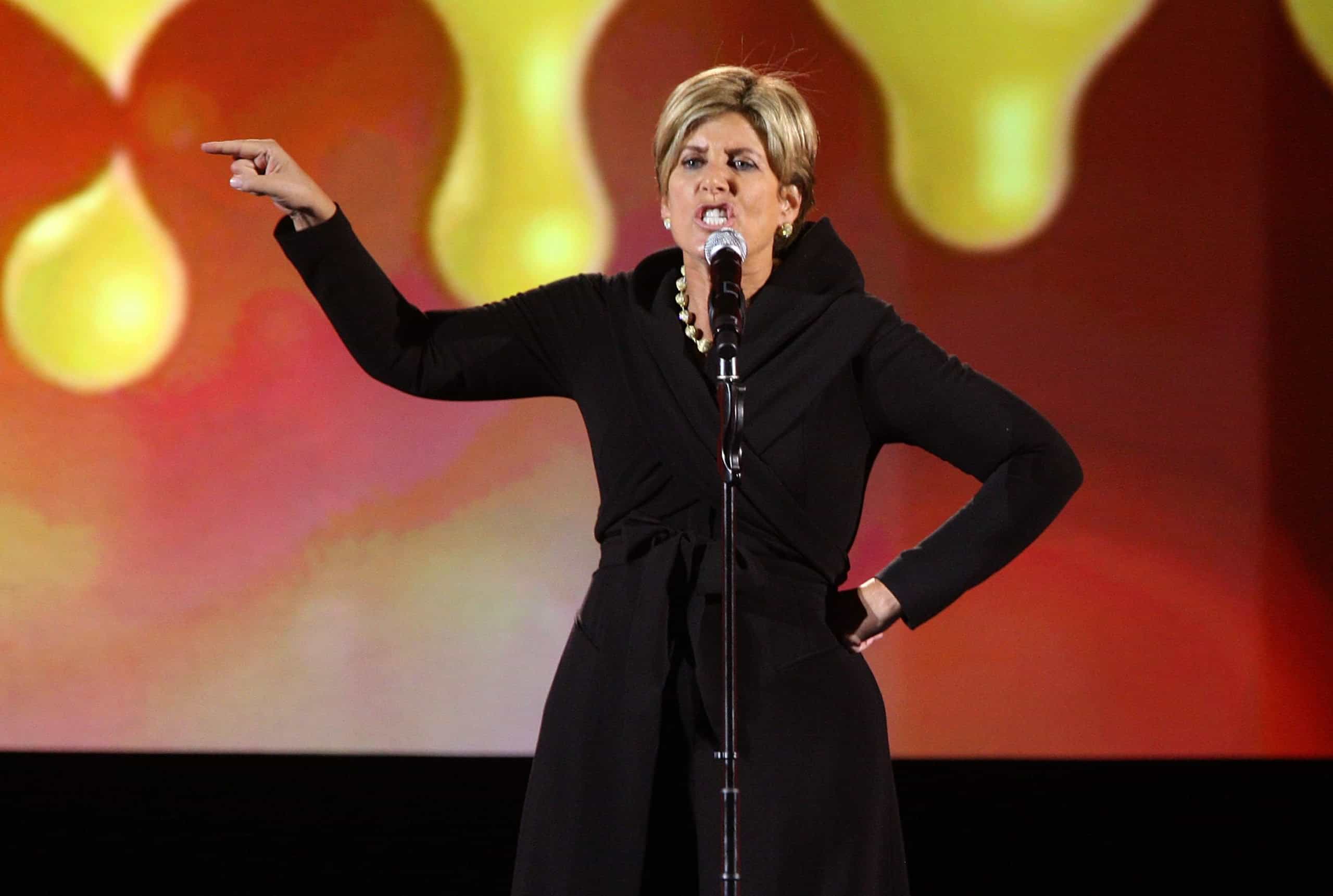 Suze Orman, a woman with short blonde hair, stands at a microphone, wearing a black long coat and a pearl necklace. She points with her left arm raised, while her right hand rests on her hip, her expression animated as she speaks. The background features blurred, glowing yellow and orange abstract shapes against a red and pink gradient.
