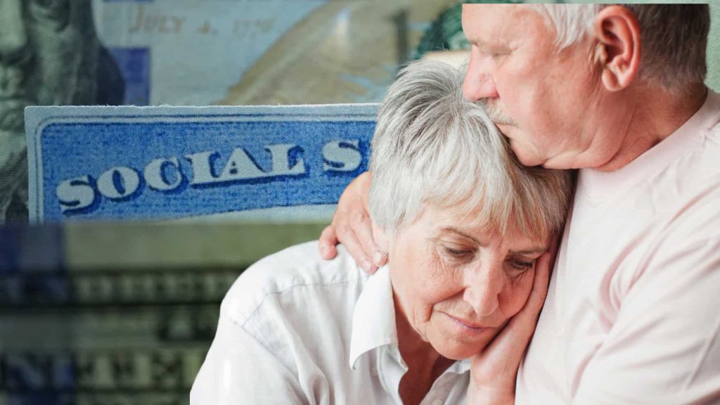 A close-up of an elderly man with a mustache comforting an elderly woman with short gray hair, who appears distressed, leaning her head on his shoulder. Her eyes are closed, and his hand is gently on her shoulder. In the blurred background, a blue document with the word 'SOCIALS' and part of a US dollar bill are visible.