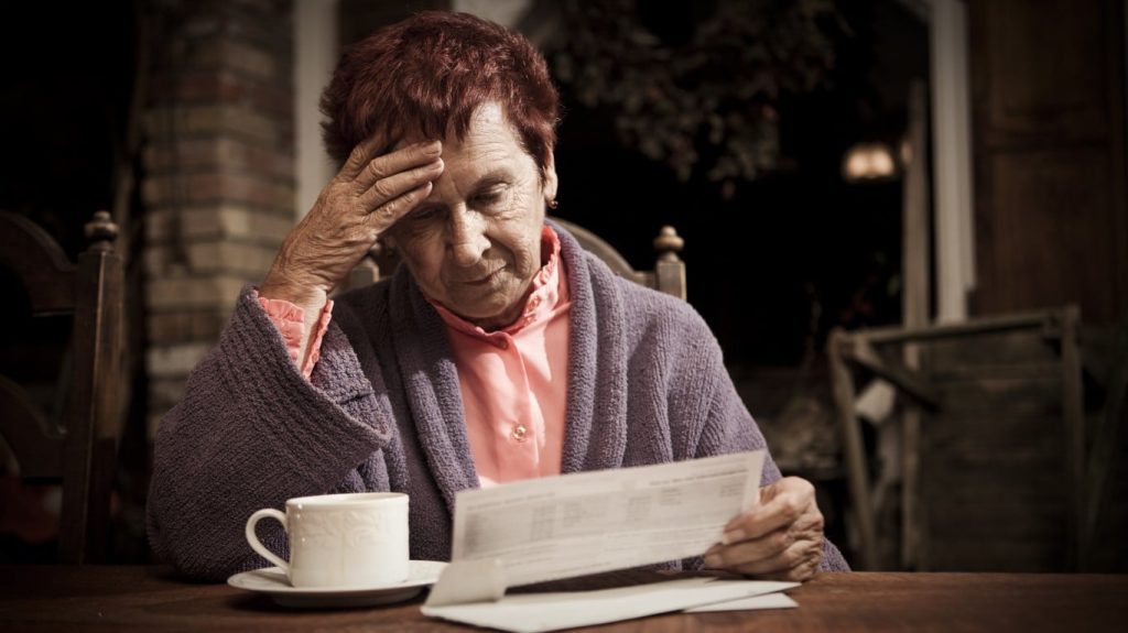 A close-up, dimly lit photo shows an elderly woman with short, reddish hair, wearing a pink shirt and a purple robe, sitting at a wooden table. She is holding a white document with both hands, looking down at it with a worried expression, her right hand resting on her forehead. A white teacup and saucer are on the table to her left. The background is dark, showing faint outlines of furniture.