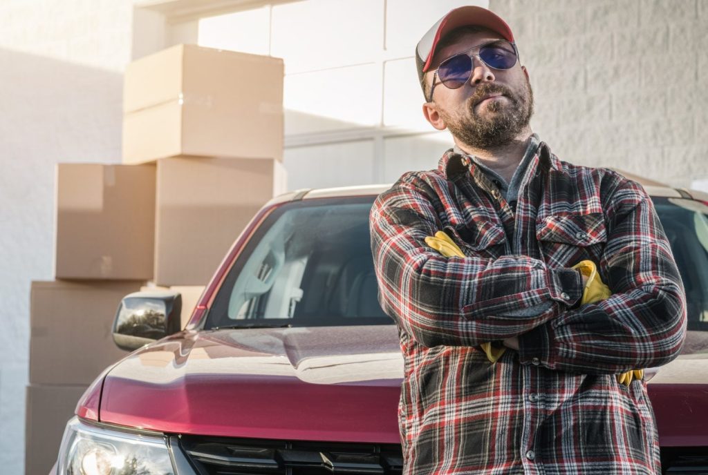 Proud Caucasian Contractor Worker in His 30s Wearing Sunglasses Next to His Modern Pickup Truck and Cargo Boxes. Supplies Delivery Theme.