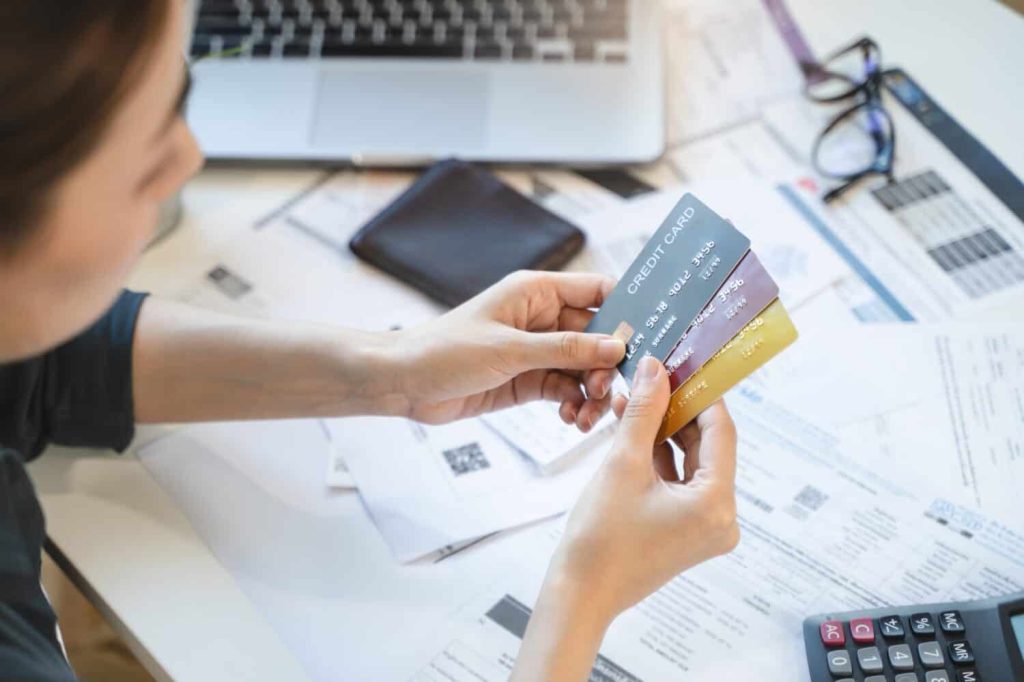 A person's hands hold three credit cards (blue, red, gold) over a desk. The desk is covered with scattered financial papers, receipts, a calculator, a laptop, and a pair of reading glasses. The person is wearing a dark shirt.
