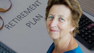 A close-up photo of an older woman with light brown hair and bright blue eyes, smiling softly while looking forward. To her left, part of a white document with the grey text 'RETIREMENT PLAN' is visible. A black scientific calculator is partially visible to her right, and the top of a pair of gold-rimmed eyeglasses is in the upper left corner. The background is predominantly light grey from the document.