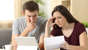 A young man in a gray t-shirt looks distressed, hand covering his mouth, while looking at a white tablet. Beside him, a young woman in a maroon top holds and reads a white letter, her face showing worry and her hand resting on her forehead. They appear to be in a living room or home office.
