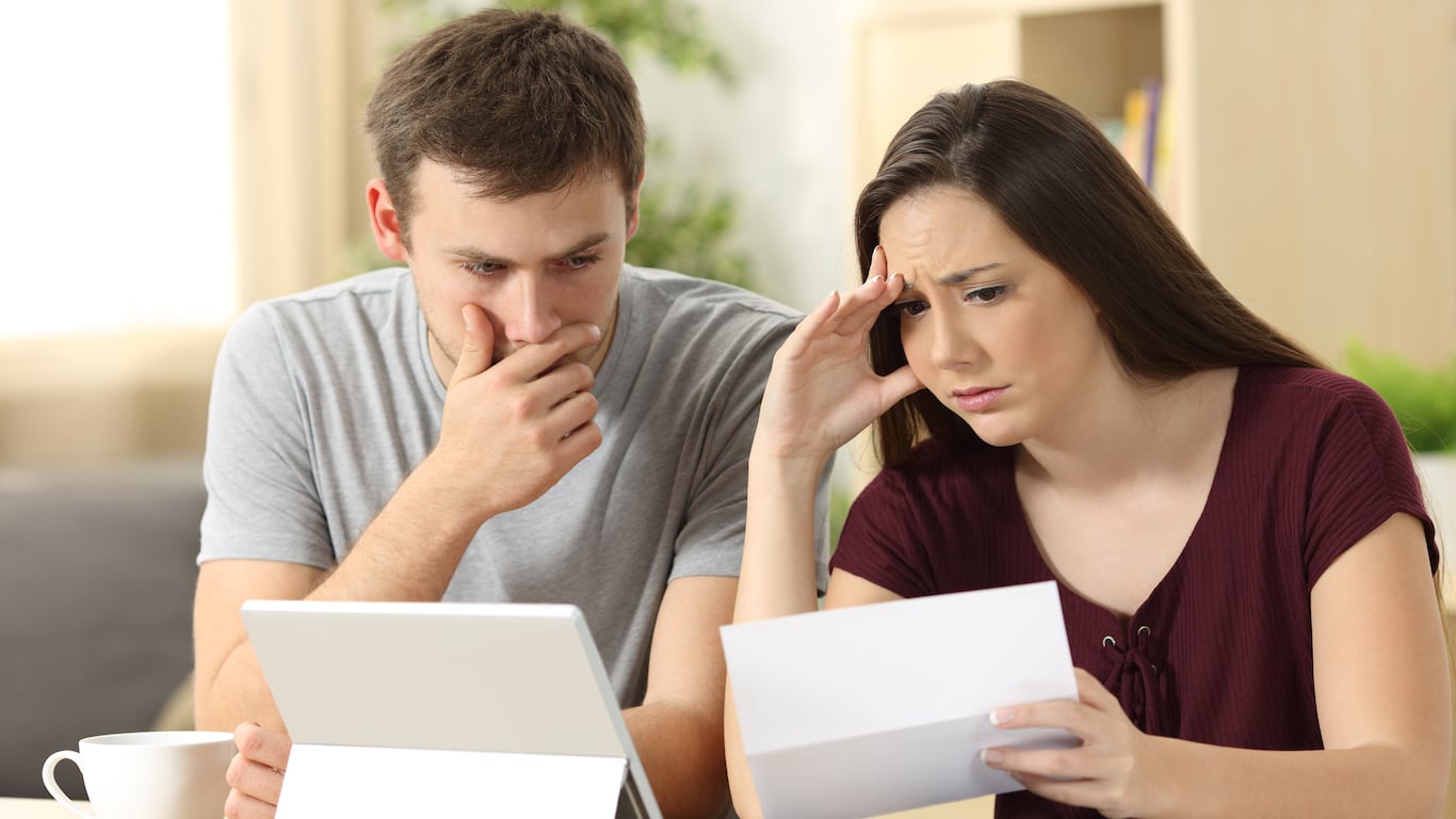A young man in a gray t-shirt looks distressed, hand covering his mouth, while looking at a white tablet. Beside him, a young woman in a maroon top holds and reads a white letter, her face showing worry and her hand resting on her forehead. They appear to be in a living room or home office.