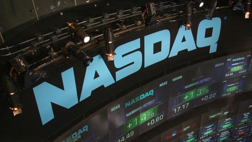 An overhead shot of the NASDAQ trading floor, featuring a large, illuminated light blue NASDAQ logo against a dark wall. Below the logo, several digital screens display stock market data with green positive percentage changes, including company names like NASDAQLISTED and Google, and tickers such as NDAQ and MBWM. Bright spotlights mounted on metal structures are visible above the sign, adding to the dramatic lighting.