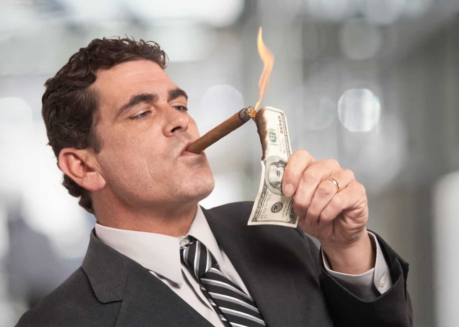 A man in a dark suit and striped tie is shown in profile, lighting a cigar held in his mouth with a burning US $100 bill. Flames rise from the top of the bill as he holds it up to the cigar. The background is a soft, out-of-focus grey.