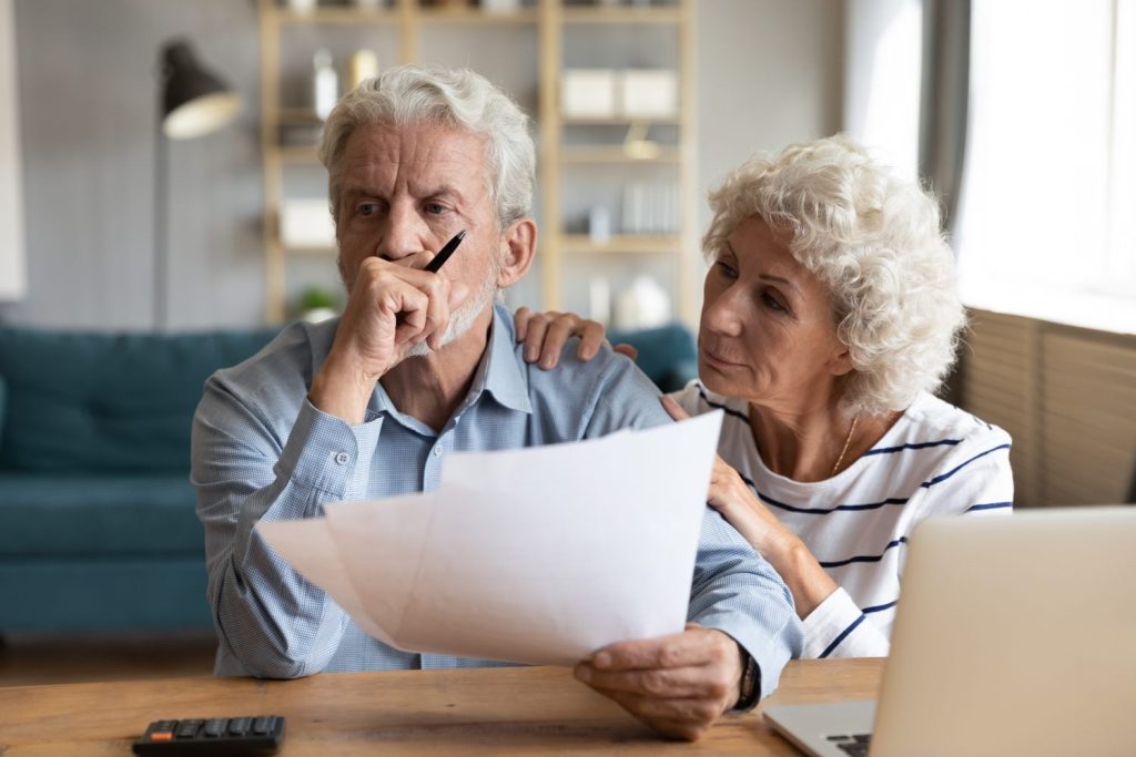 A close-up of an elderly couple at a wooden table. The man, with gray hair and a beard, wears a light blue patterned shirt, holds a pen to his chin, and looks concerned while holding white papers. The woman, with curly white hair, wears a white and blue striped shirt and has her hand on the man's shoulder, looking thoughtfully in the same direction. A black calculator and part of a silver laptop are visible on the table. The background shows a softly lit living room.