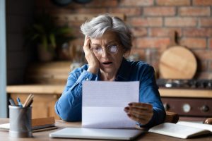 An older woman with short gray hair and round glasses sits at a wooden table, holding a white letter. Her expression is one of shock and distress, with her mouth slightly open and one hand touching her face. A laptop, an open book, and a pen holder are also visible on the table. The background features a brick wall and kitchen elements.