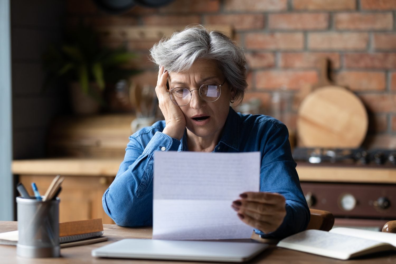 An older woman with short gray hair and round glasses sits at a wooden table, holding a white letter. Her expression is one of shock and distress, with her mouth slightly open and one hand touching her face. A laptop, an open book, and a pen holder are also visible on the table. The background features a brick wall and kitchen elements.