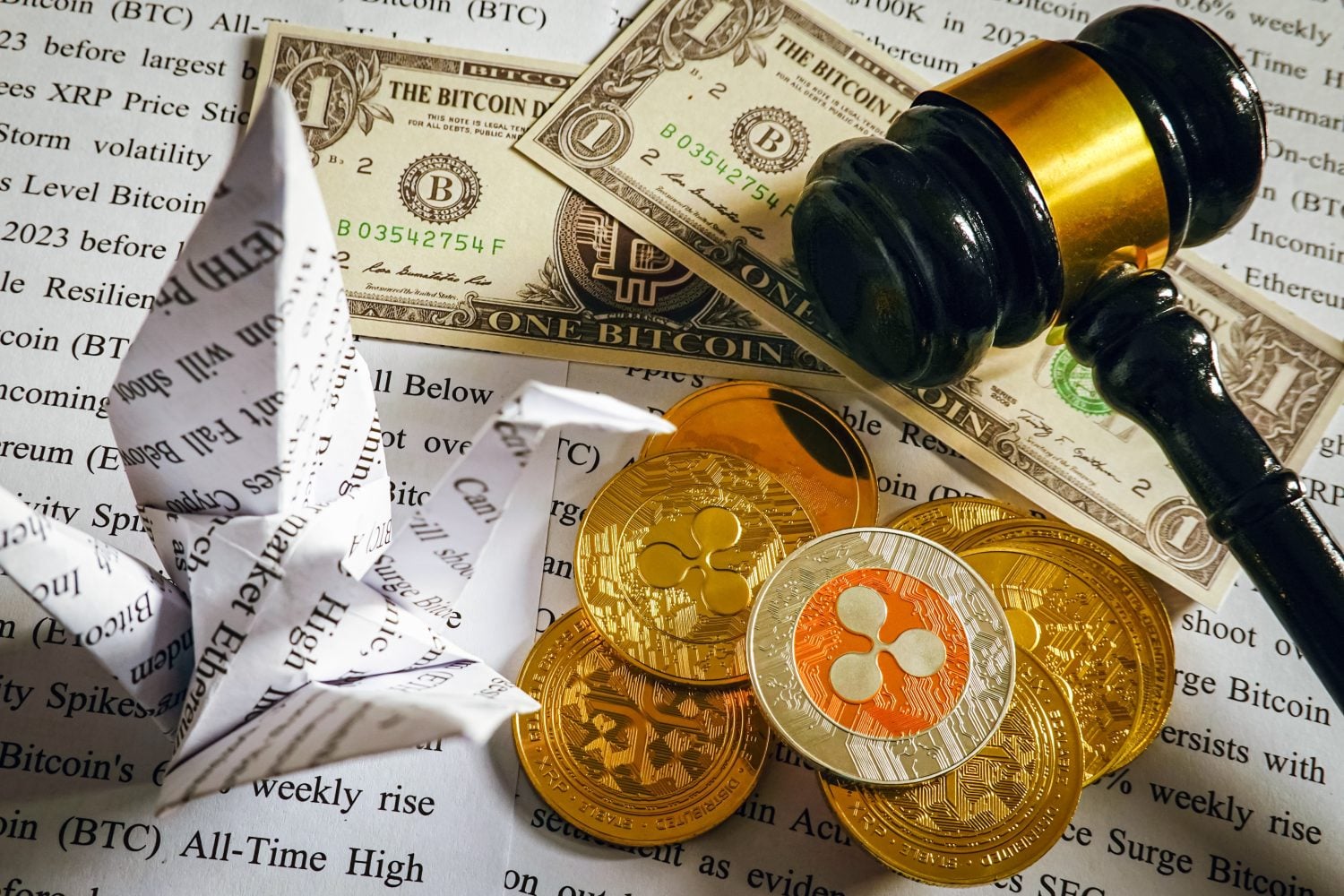 A top-down view shows a black and gold wooden gavel resting on a stack of gold cryptocurrency coins and US dollar bills, one bill featuring 'THE BITCOIN D' text. A prominent silver and orange Ripple coin lies on top of the gold coins. An origami crane, folded from newspaper text, is on the left, with blurred financial article text forming the background.