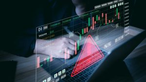 A close-up, dimly lit photograph shows a person's hands resting on a laptop keyboard. A translucent, holographic display of financial market data, featuring green and red candlestick charts, numerical values, and a grid, is overlaid above the laptop. A large, bright red triangular warning symbol with an exclamation mark is prominently displayed over the laptop keyboard and the lower part of the screen, creating a serious and cautious mood related to financial analysis.