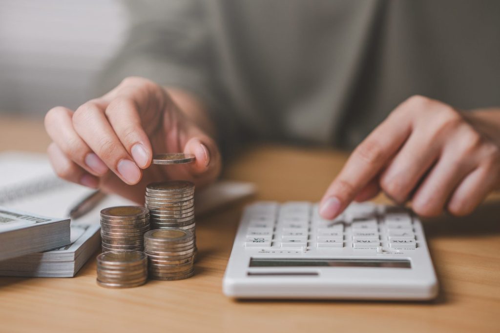 Close-up of a person's hands at a light wooden desk. The left hand carefully places a silver coin onto a stack of similar coins, with three other coin stacks of varying heights nearby. The right hand presses buttons on a white calculator. In the background, a stack of hundred-dollar bills and a spiral-bound notebook with a pen are visible, creating a focused financial scene.