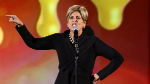 Suze Orman, a woman with short blonde hair, stands at a microphone, wearing a black long coat and a pearl necklace. She points with her left arm raised, while her right hand rests on her hip, her expression animated as she speaks. The background features blurred, glowing yellow and orange abstract shapes against a red and pink gradient.