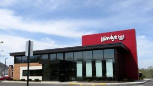 A modern Wendy's fast-food restaurant building with a large red section on the right displaying the white 'Wendy's' logo and the cartoon character. The building features dark framed windows, a dark roofline, and sections of brick and white paneling. A light blue sky with wispy clouds is visible above. A black and white signpost is in the foreground, and a parking lot with yellow lines is visible at the bottom.