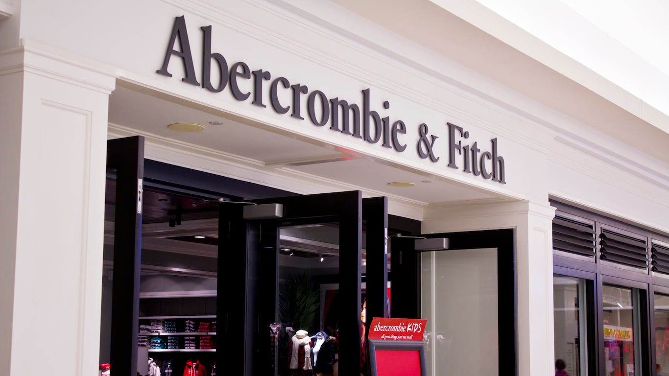 A straight-on view of the Abercrombie & Fitch store entrance. The store name in dark, large, serif letters is mounted on a white architectural facade above dark-framed glass double doors. Inside, some clothing racks are visible. A small red sign for 'Abercrombie KIDS' stands near the right door.