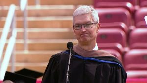 Tim Cook stands at a podium microphone wearing black academic regalia with a blue hood stripe, speaking at a university commencement ceremony with red auditorium seating visible behind him.