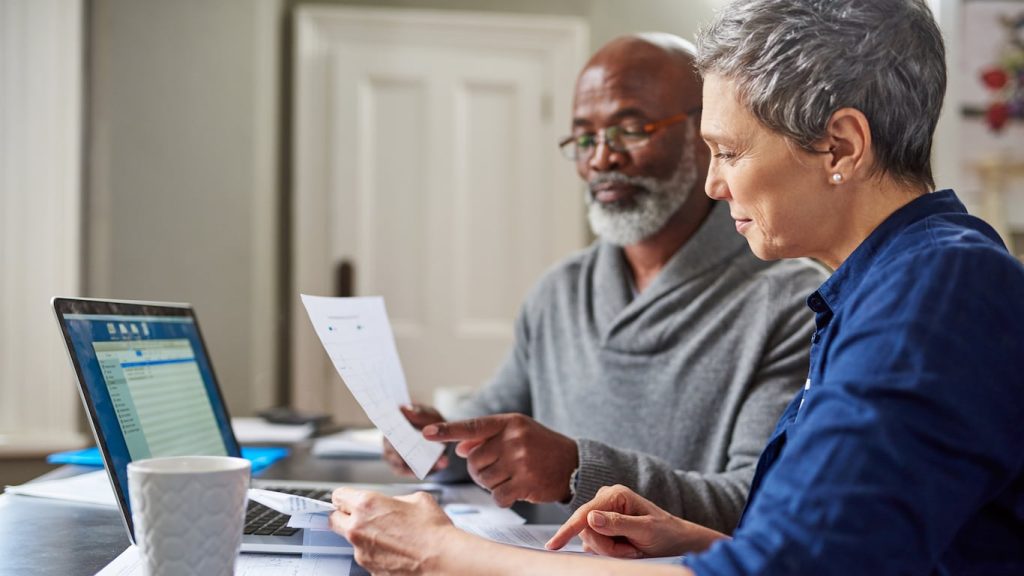 A diverse older couple sits at a table, both focused on paperwork and a silver laptop. The man, with a gray beard and glasses, wears a gray sweater and points at a document he holds. The woman, with short gray hair and a blue button-up shirt, holds papers and also points at a document on the table, with a white coffee mug visible near the laptop.