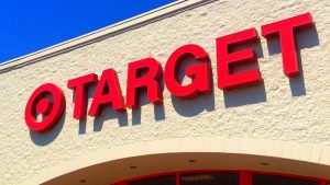A low-angle close-up shot of the red, three-dimensional 'TARGET' store sign mounted on a light beige, textured brick wall under a bright blue sky. The letters cast distinct shadows on the wall, and a curved architectural element is visible below the sign.