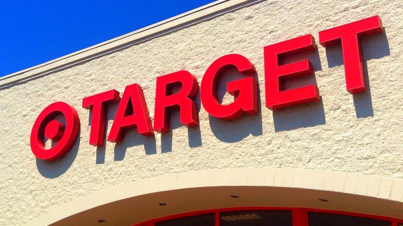 A low-angle close-up shot of the red, three-dimensional 'TARGET' store sign mounted on a light beige, textured brick wall under a bright blue sky. The letters cast distinct shadows on the wall, and a curved architectural element is visible below the sign.
