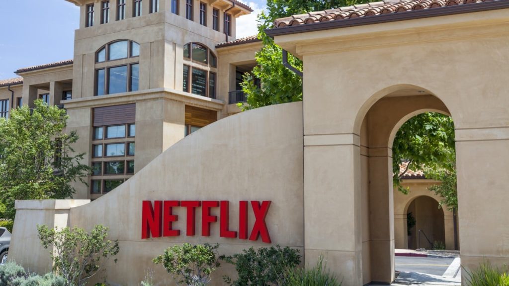 A wide, sunlit exterior shot of the Netflix corporate campus. In the foreground, a curved, light beige stucco wall prominently displays the red, sans-serif 'NETFLIX' logo. Behind the wall, a modern building with multiple windows and balconies is visible. To the right, an arched entryway leads to another part of the campus. Green trees and shrubs are scattered around the base of the wall and buildings under a bright blue sky.