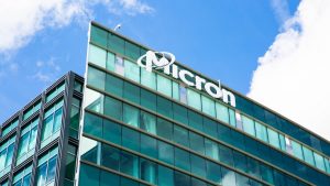 A low-angle exterior shot of a modern glass-paneled building with the white 'Micron' logo clearly visible on its facade. The building has a green-blue reflective glass exterior under a bright blue sky with white clouds. A security camera is mounted on the top edge of the building.