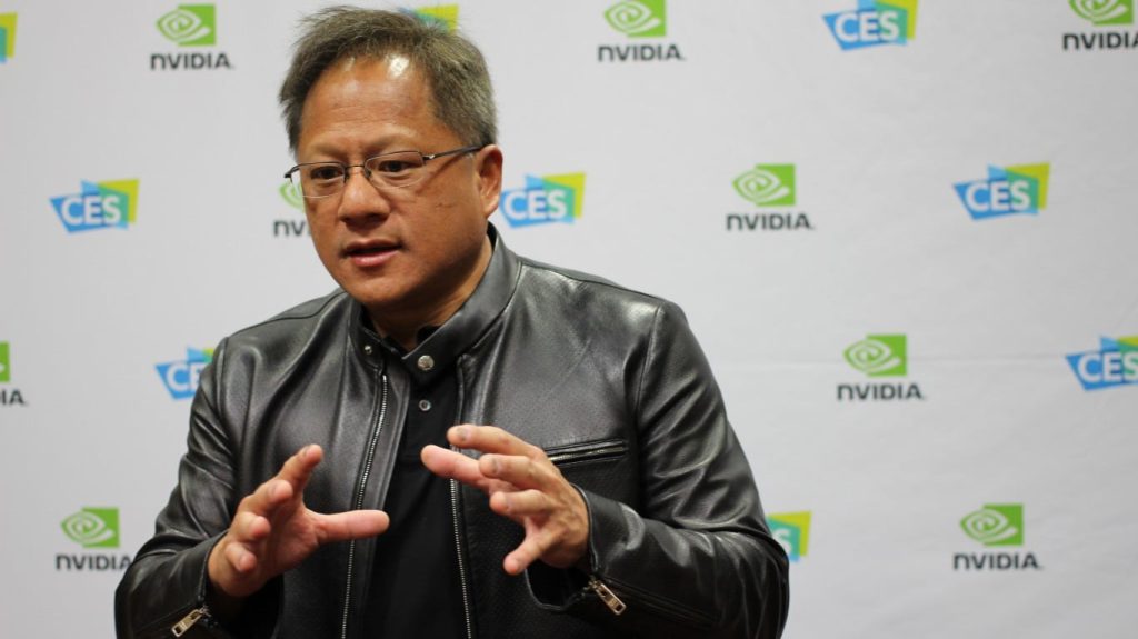 A medium shot of Nvidia CEO Jensen Huang, an East Asian man, wearing glasses and a black leather jacket. He is looking slightly to his right, speaking and gesturing with both hands. The background is a white wall with repeating green NVIDIA logos and blue CES logos.