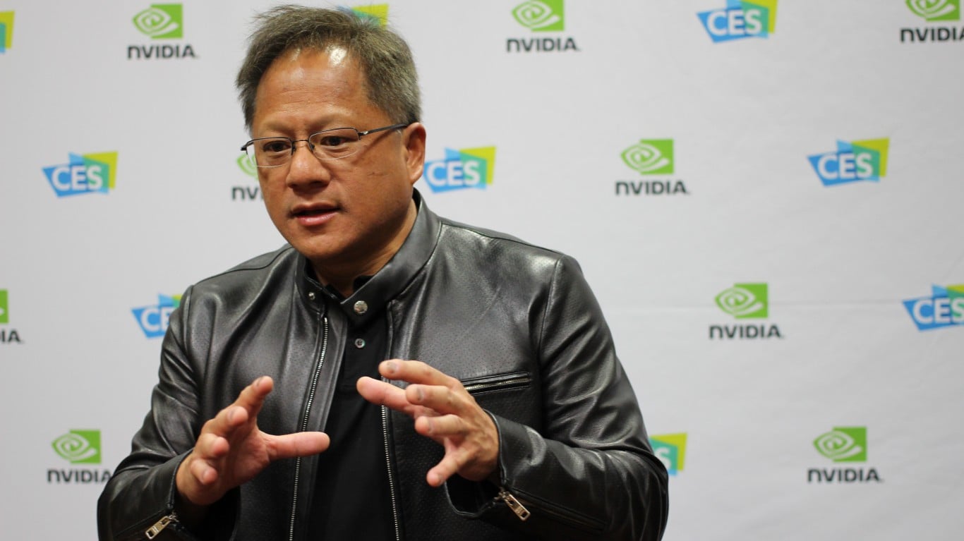 A medium shot of Nvidia CEO Jensen Huang, an East Asian man, wearing glasses and a black leather jacket. He is looking slightly to his right, speaking and gesturing with both hands. The background is a white wall with repeating green NVIDIA logos and blue CES logos.