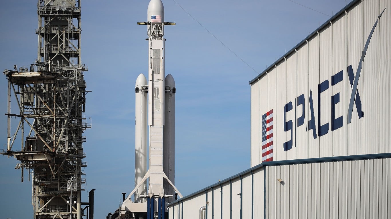 A tall, white SpaceX Falcon Heavy rocket stands on a launchpad next to a large, intricate metal scaffolding structure. To the right of the rocket, part of a white industrial building with the blue 'SpaceX' logo is visible, featuring red and white stripes on the letter 'X'. An American flag is visible near the top of the rocket.