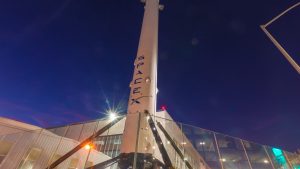 A tall white SpaceX Falcon 9 rocket, featuring the 'SPACE X' logo vertically in blue, stands against a deep blue night sky. Bright, star-like lens flares from unseen lights illuminate the scene, with portions of a modern building and a glass barrier visible around the rocket's base.