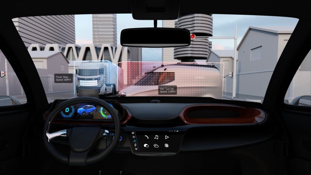 View from inside a car with a black steering wheel and dark interior, looking out through the windshield onto a city street. An augmented reality overlay displays digital information: a blue outlined truck to the left labeled 'Truck: Stop Speed: 0MPH' and a red outlined van to the right labeled 'Van: Turn left Speed: 5 MPH'. Buildings and generic city infrastructure are visible through the windshield. The dashboard includes a digital screen showing a blue SUV graphic and a speed gauge at '0'. Below, a secondary touchscreen displays various app icons such as music, calls, and weather.