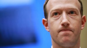 A close-up portrait of Mark Zuckerberg showing his face from the eyes down to just below the chin. He has fair skin with visible pores and subtle stubble. His light blue eyes gaze slightly upwards, and his lips are pressed together in a neutral, slightly downturned expression. The background is a soft, out-of-focus blue.