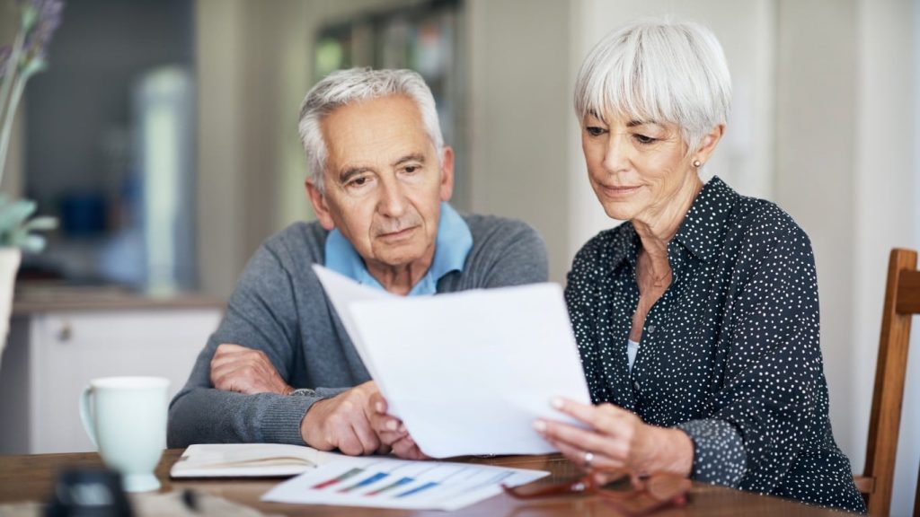 An older white couple sits at a wooden table, both intently looking at white papers held by the woman. The man, with short grey hair and a blue polo shirt under a grey cardigan, leans in from the left, looking serious. The woman, with short grey hair and a black shirt with white polka dots, holds the documents. On the table, a light blue mug, an open notebook with colorful bar charts, and some eyeglasses are visible. The background shows a blurred, bright home interior.
