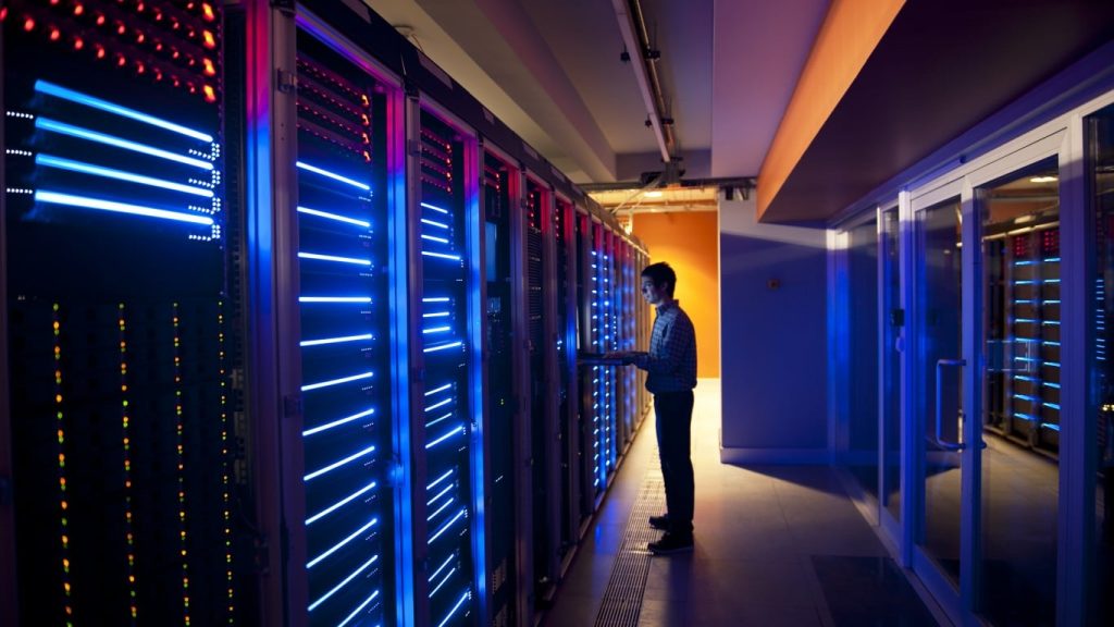 A person in a plaid shirt stands in a server room, looking at a laptop or tablet. They are surrounded by rows of tall server racks that emit glowing blue and red lights. The floor has grates, and there's a door with glass panels on the right, leading to another section of the facility. The lighting is dark with strong colorful illumination from the servers.