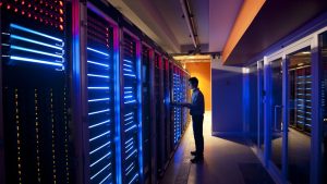 A person in a plaid shirt stands in a server room, looking at a laptop or tablet. They are surrounded by rows of tall server racks that emit glowing blue and red lights. The floor has grates, and there's a door with glass panels on the right, leading to another section of the facility. The lighting is dark with strong colorful illumination from the servers.