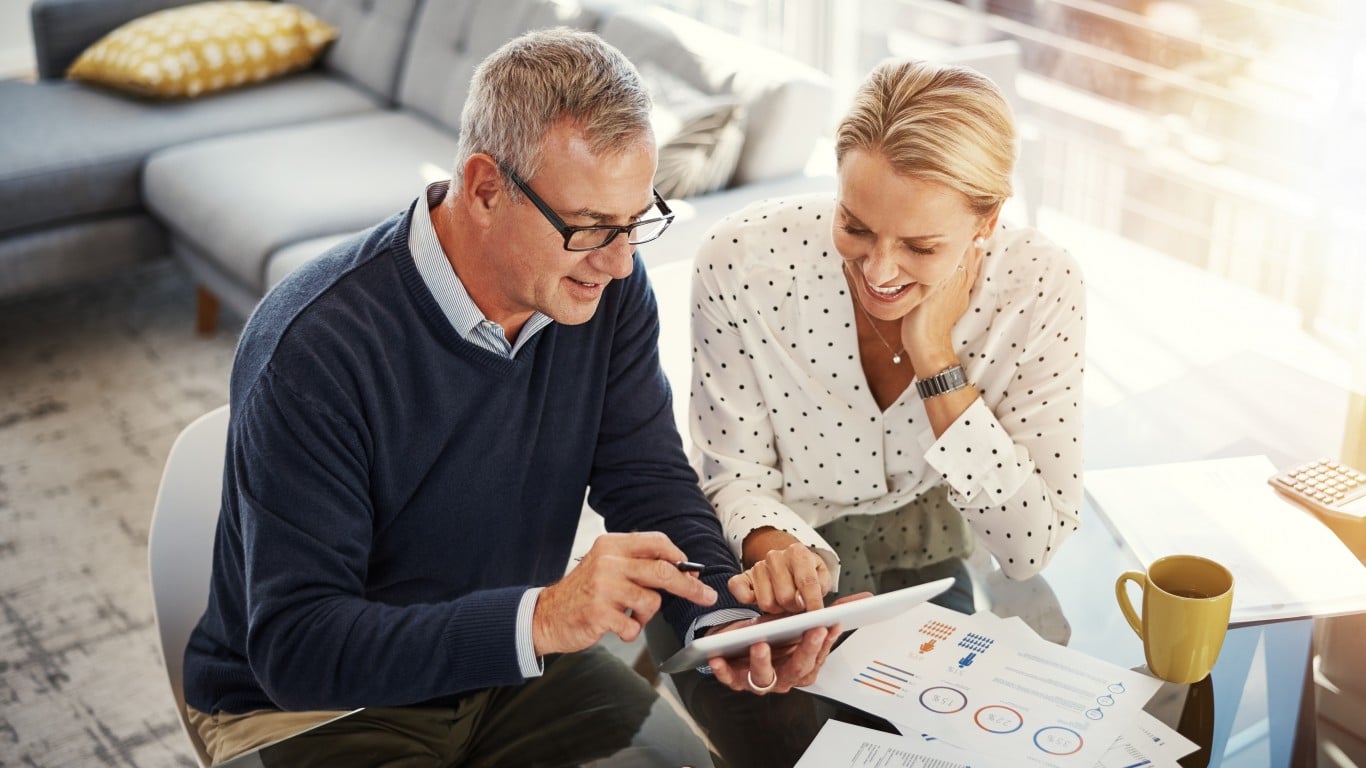 A man wearing glasses and a navy blue sweater points to a white tablet screen, while a blonde woman in a white polka-dot blouse smiles and looks at the screen. They are seated at a glass table with several financial charts and graphs on papers, a yellow mug, and a calculator. A gray sectional couch and a sunlit window are visible in the background, creating a bright and comfortable home or office setting.