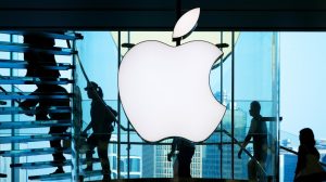 A large, glowing white Apple logo is prominently displayed in a modern building with blue-tinted glass walls and staircases. Silhouetted people walk on the transparent stairs and past the logo, with light blue skies and distant city buildings visible through the glass. The scene has a sleek, high-tech, and dynamic feel.