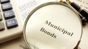 A magnifying glass is placed on a document, enlarging the bold text 'Municipal Bonds'. To the left, a white calculator with blue and gray buttons ('MR', 'M-', 'M+') is visible. In the background, a financial spreadsheet with rows of numbers like '4581.21' and '7825.66' can be seen, along with a gold and black fountain pen at the top right.