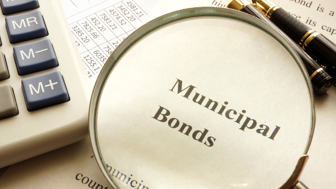 A magnifying glass is placed on a document, enlarging the bold text 'Municipal Bonds'. To the left, a white calculator with blue and gray buttons ('MR', 'M-', 'M+') is visible. In the background, a financial spreadsheet with rows of numbers like '4581.21' and '7825.66' can be seen, along with a gold and black fountain pen at the top right.