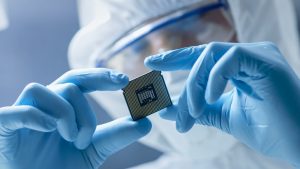 A close-up shows a person's hands, covered in bright blue latex gloves, holding a dark square computer microchip between their thumb and forefinger. The chip displays a dense grid of small gold pins on its underside and a rectangular array of metallic contacts in its center. In the blurred background, parts of the person's white protective cleanroom suit and blue safety goggles are visible, indicating a sterile laboratory setting under bright, even lighting.