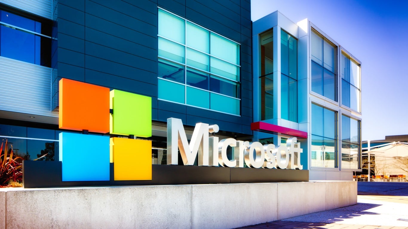 A wide shot of the Microsoft corporate campus on a sunny day. A large, silver 'Microsoft' logo is displayed on a low concrete wall in front of a modern building. Above the text logo are four brightly colored squares: orange, lime green, bright blue, and yellow, forming the iconic Microsoft symbol. The building behind features dark paneling and reflective blue windows, with a sharp, modern architectural style.