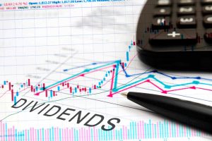 A close-up view of a financial chart on white paper, displaying colorful stock market data with red and blue candlestick patterns and wavy line graphs. The word 'DIVIDENDS' is written in large, bold letters across the lower left section. A black calculator is partially visible in the upper right corner, and a black pen with a gold tip rests on the paper, pointing towards the financial information.