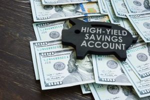 A black wooden piggy bank-shaped sign with 'HIGH-YIELD SAVINGS ACCOUNT' written in white capital letters is placed on a scattered pile of US hundred-dollar bills, all resting on a dark brown wooden table.