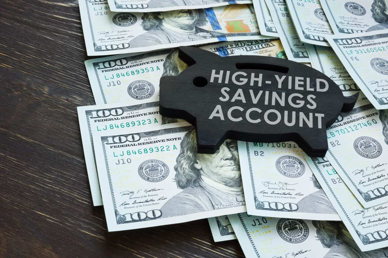 A black wooden piggy bank-shaped sign with 'HIGH-YIELD SAVINGS ACCOUNT' written in white capital letters is placed on a scattered pile of US hundred-dollar bills, all resting on a dark brown wooden table.
