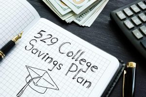 A black and gold fountain pen rests on an open, grid-lined notebook. The notebook displays handwritten text '529 College Savings Plan' and a line drawing of a graduation cap. In the background, stacks of US hundred-dollar bills and a dark gray calculator are visible on a dark wood desk.