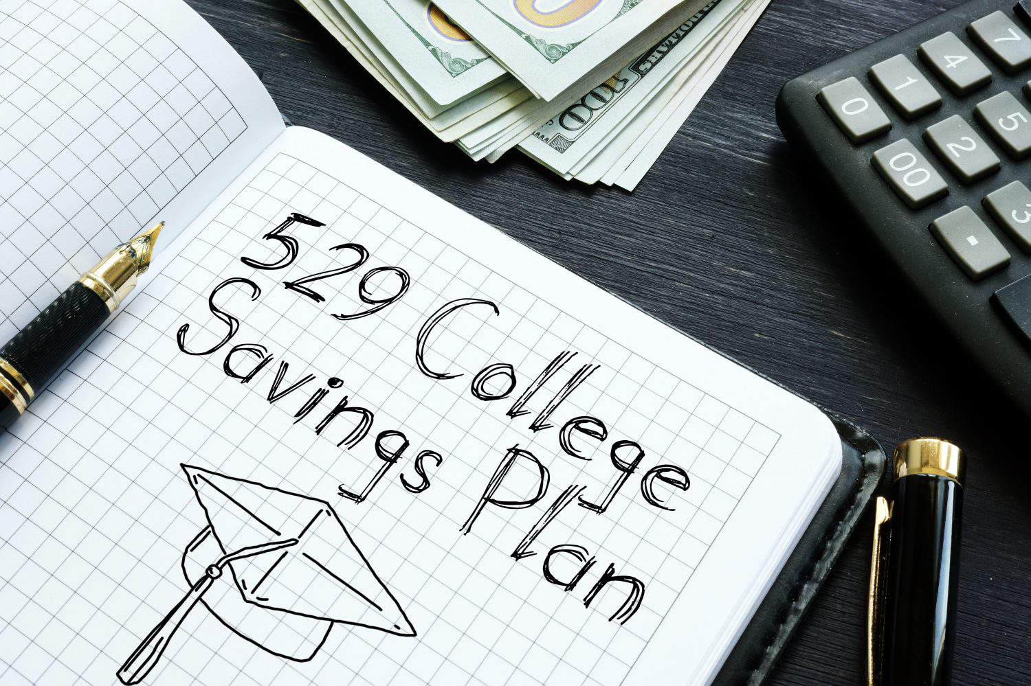 A black and gold fountain pen rests on an open, grid-lined notebook. The notebook displays handwritten text '529 College Savings Plan' and a line drawing of a graduation cap. In the background, stacks of US hundred-dollar bills and a dark gray calculator are visible on a dark wood desk.