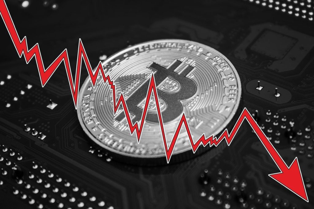 A silver Bitcoin coin rests on a dark gray or black electronic circuit board. A bright red, jagged line with a white outline plots a sharp downward trend from the upper left to the lower right of the image, ending in a prominent red arrow pointing down. The red line zigzags across the surface of the Bitcoin coin and the circuit board, visually representing a market decline.