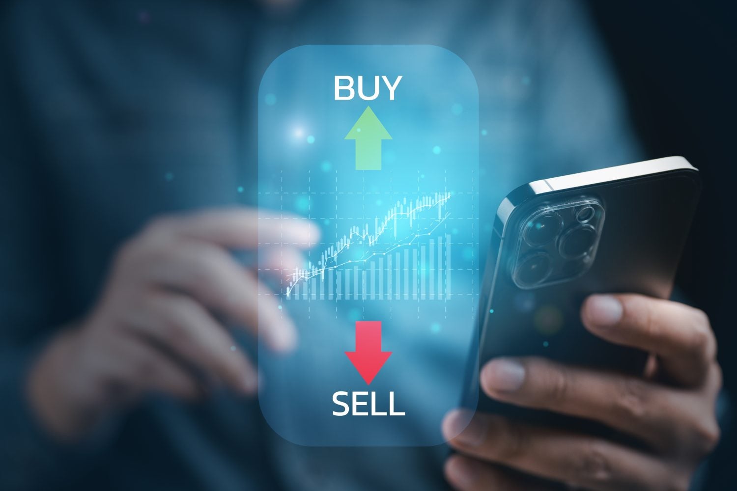 A close-up shot of a person's hands holding a dark grey smartphone. Overlaid on the scene is a translucent blue rectangular screen displaying a financial graph with both line and bar charts against a grid. Above the graph is a large green upward arrow with the word 'BUY', and below it is a large red downward arrow with the word 'SELL'. The background is blurred, focusing on the hands, phone, and digital interface.