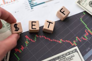 A person's hand places a wooden block with a dollar sign ($) next to other wooden blocks that spell out 'ETF' on a black financial chart. The chart displays red and green candlestick patterns, indicating market movements. US dollar bills and a blurred financial report are visible in the background.